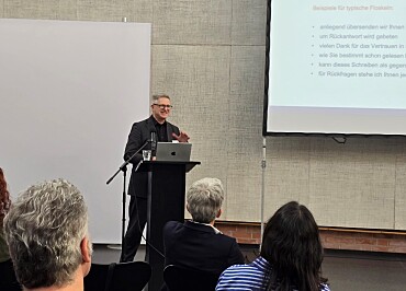 Im Vordergrund sieht man Publikum von hinten. Im Hintergrund steht Professor Brettschneider am Redepult. Rechtsoberhalb von ihm ist eine Präsentationsfolie mit der Überschrift Vermeiden Sie Floskeln! zu sehen.