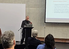 Im Vordergrund sieht man Publikum von hinten. Im Hintergrund steht Professor Brettschneider am Redepult. Rechtsoberhalb von ihm ist eine Präsentationsfolie mit der Überschrift Vermeiden Sie Floskeln! zu sehen.