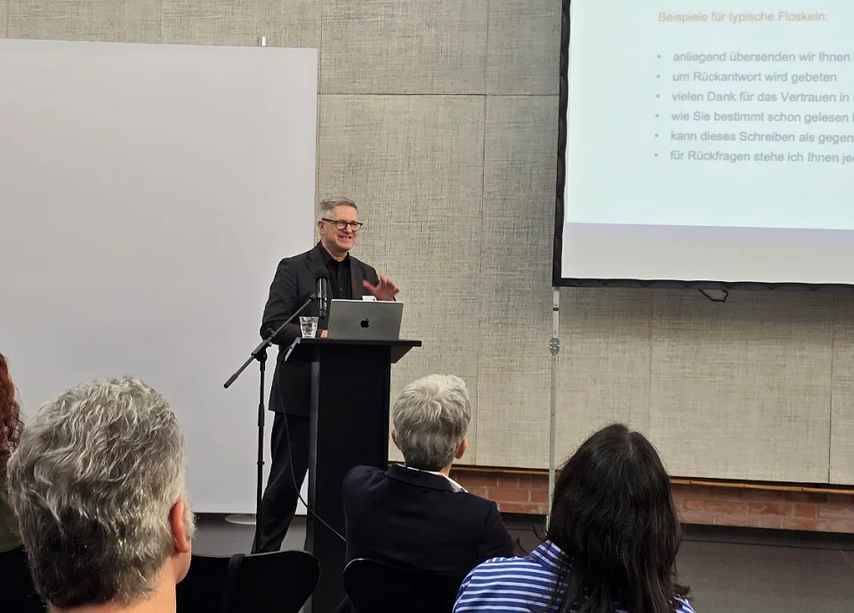 Im Vordergrund sieht man Publikum von hinten. Im Hintergrund steht Professor Brettschneider am Redepult. Rechtsoberhalb von ihm ist eine Präsentationsfolie mit der Überschrift Vermeiden Sie Floskeln! zu sehen.