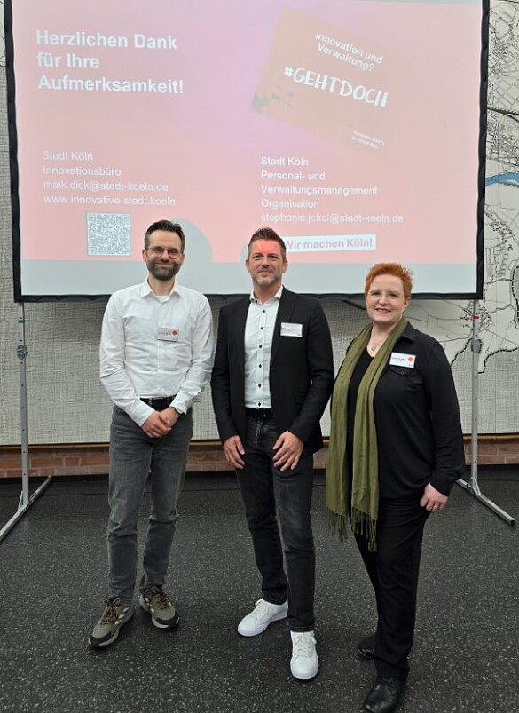 Drei Personen stehen nebeneinander und sehen in die Kamera: Dr. David Sörgel, Maik Dick, Stephanie Jekel (v. l. n. r.) Dahinter ist eine Präsentationsfolie zu sehen mit Kontaktdaten vom Innovationsbüro und vom Personal- und Verwaltungsmanagement der Stadt Köln