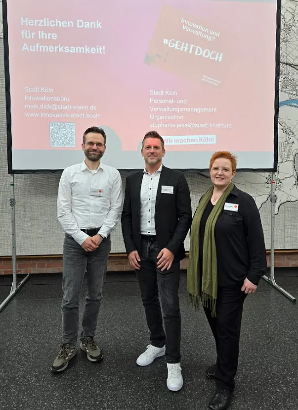 Drei Personen stehen nebeneinander und sehen in die Kamera: Dr. David Sörgel, Maik Dick, Stephanie Jekel (v. l. n. r.) Dahinter ist eine Präsentationsfolie zu sehen mit Kontaktdaten vom Innovationsbüro und vom Personal- und Verwaltungsmanagement der Stadt Köln