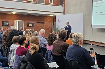 Im Bildhintergrund steht Bremens Finanzsenator Björn Fecker an einem Redepult, im Vordergrund ist das Publikum von schräg hinten zu sehen