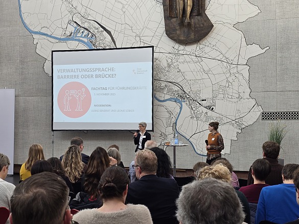 Das Foto zeigt die Moderatorinnen, Ulrike Bendrat und Leonie Lübker, vor dem Fachtagspublikum aus Führungskräften in der Lichthofhalle des Veranstaltungsortes Kwadrat. Im Hintergrund ist die Präsentation auf einer Leinwand zu sehen.
