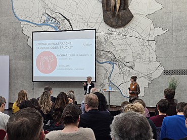 Das Foto zeigt die Moderatorinnen, Ulrike Bendrat und Leonie Lübker, vor dem Fachtagspublikum aus Führungskräften in der Lichthofhalle des Veranstaltungsortes Kwadrat. Im Hintergrund ist die Präsentation auf einer Leinwand zu sehen.