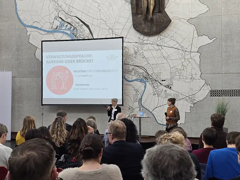 Das Foto zeigt die Moderatorinnen, Ulrike Bendrat und Leonie Lübker, vor dem Fachtagspublikum aus Führungskräften in der Lichthofhalle des Veranstaltungsortes Kwadrat. Im Hintergrund ist die Präsentation auf einer Leinwand zu sehen.